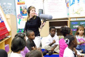Woman holding large microphone in a classroom, surrounded by young children